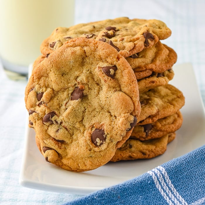 The Best Chocolate Chip Cookies photo of stacked cookies on a white plate with a glass of milk in the background