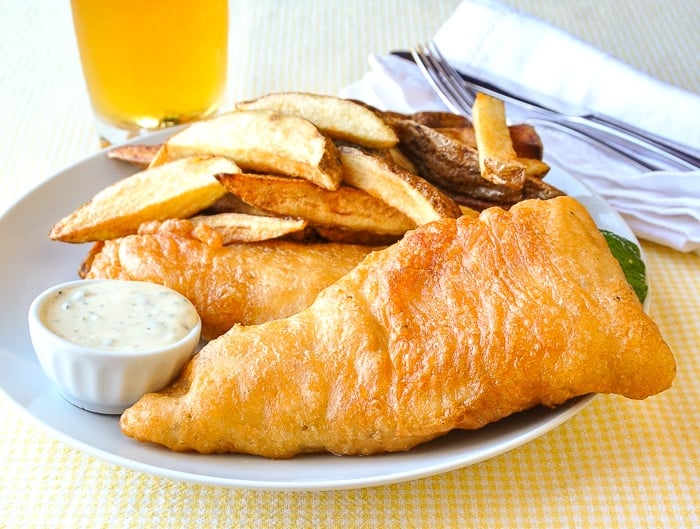 Fish and chips shown with tartar sauce and a glass of beer in the background