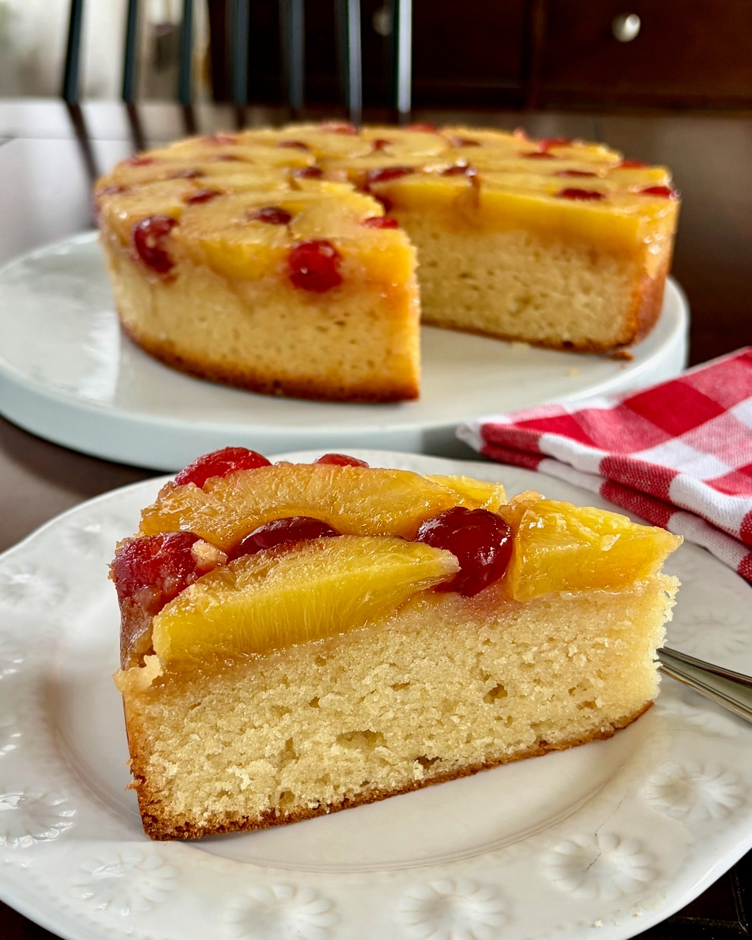 Photo of one slice of Pineapple Upside Down Cake on a white plate with whole cake in the background.