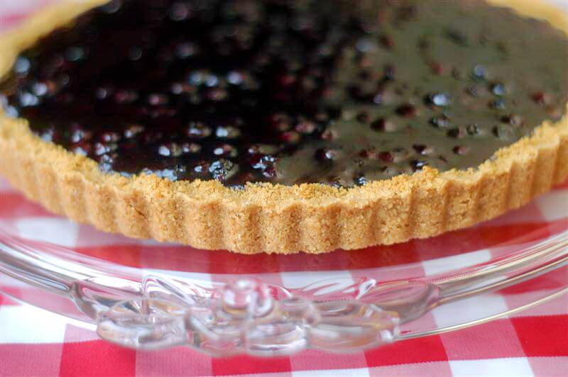 Photo of an uncut Lemon Blueberry Tart on a clear glass rose embossed cake plate.
