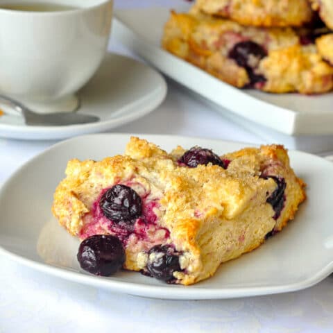 Cherry Cheesecake Scones. Single scone on plate with cup of tea and platter of scones in background