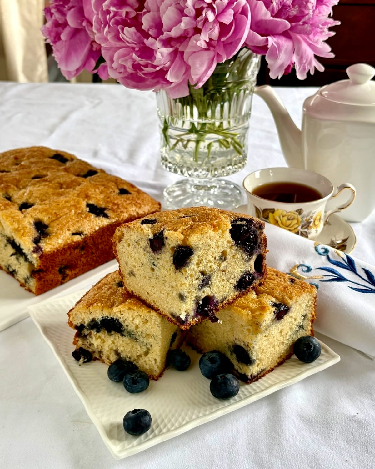 Photo of pieces of blueberry snack cake on a white serving platter with tea service and peony flowers in the background