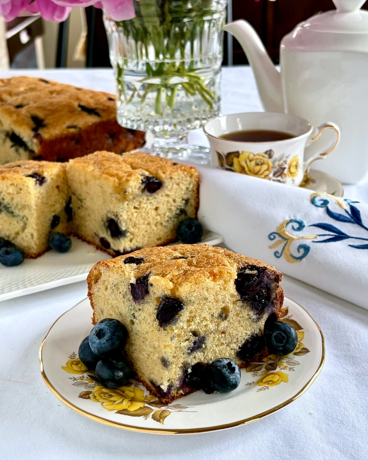Photo of one slice of blueberry snack cake with full cake in the background and tea service with napkin