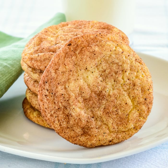 The Best Snickerdoodles Recipe close up photo of cookies stacked on a white plate with a glass of milk in the background