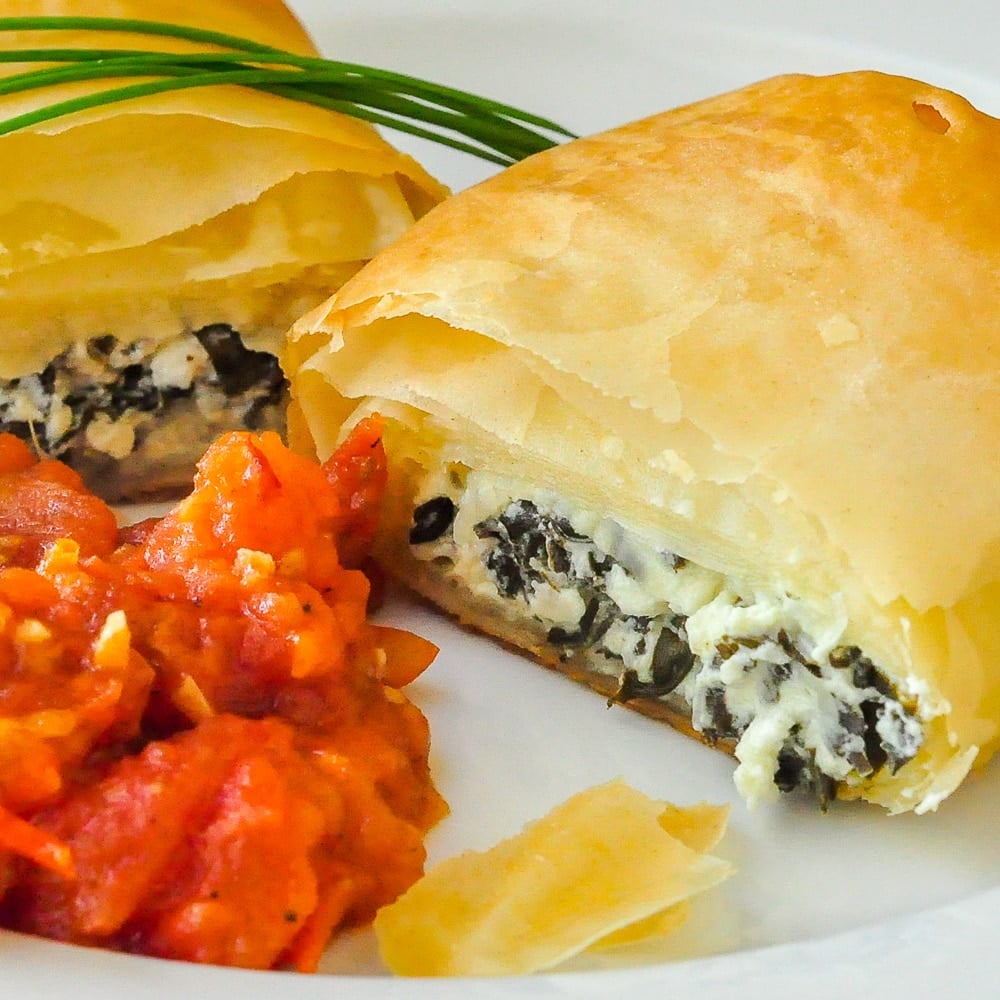 https://www.rockrecipes.com/wp-content/uploads/2012/10/Goat-Cheese-Spanakopita-Rolls-close-up-shot-of-single-roll-with-tomato-compote-on-a-white-plate.jpg