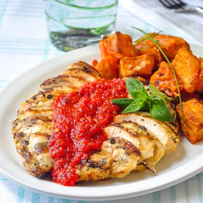 Lemon Herb Grilled Chicken with Spicy Roasted Red Pepper Sauce shown with roasted sweet potatoes on a white plate.