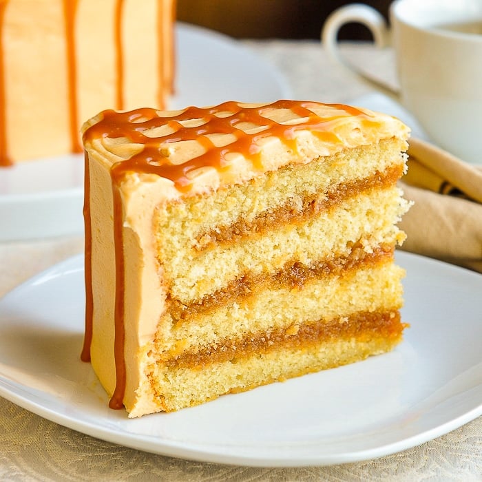 The Best Caramel Cake slice of cake on a white plate with coffee in background