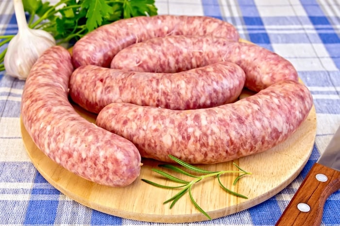 Raw pork sausages on a round wooden board, knife, rosemary, parsley and garlic on blue background checkered cloth. Licensed stock photo