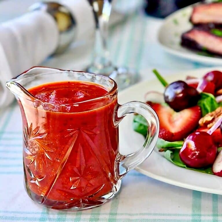 Roasted Red Pepper Salad Dressing. Photo of dressing in a clear glass jug with mixed salad on a plate beside.