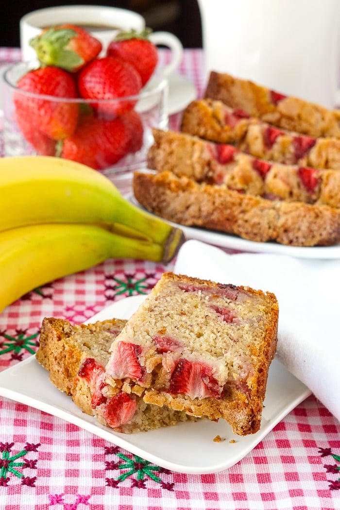 Strawberry Banana Bread photo of a single slice on white plate with coffee, bananas and strawberries in background