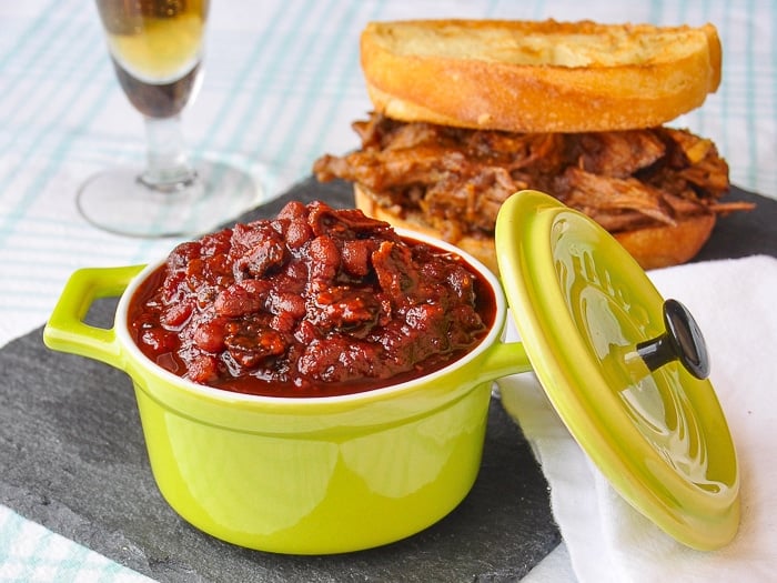 Old Fashioned Molasses and Bacon Baked Beans in a green serving dish with a pulled pork sandwich in the background