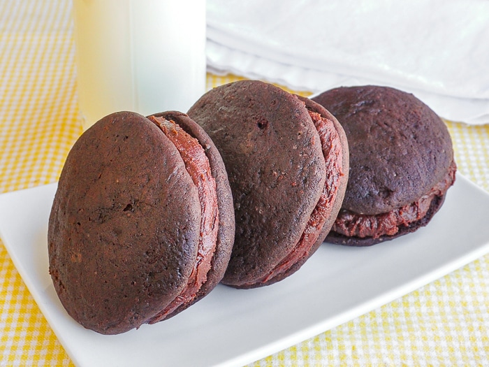 Three double chocolate whoopie pies on a white plate with a glass of milk in background