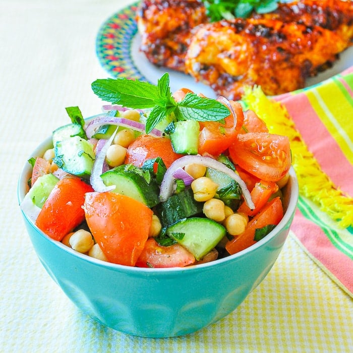 Tomato Cucumber Salad with Chickpeas & Mint shown in blue bowl with BBQ chicken in the background