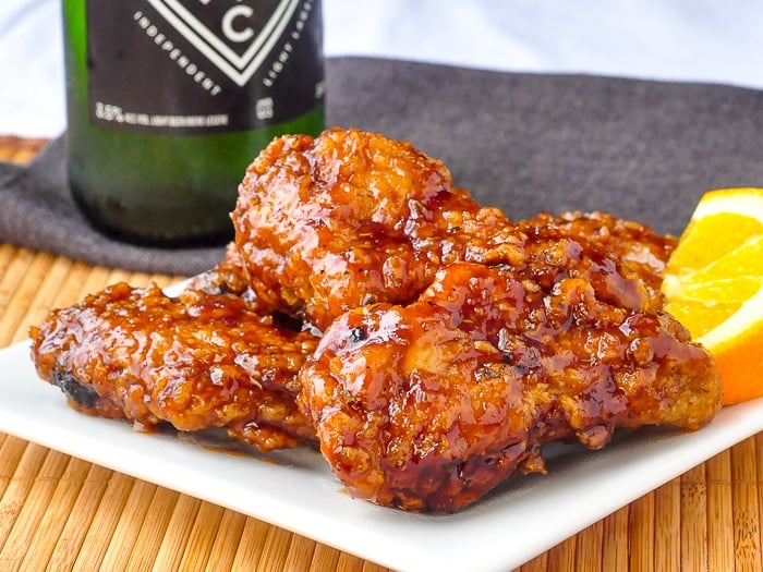 Crispy Orange Ginger Wings stacked on a white plate with a bottle of beer in the background