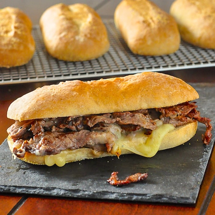 Homemade Philly Cheesesteak sandwich on a piece of stone slate with fresh rolls in background.