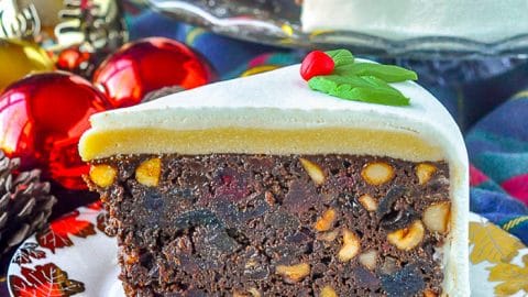 Ultimate Nut Fruitcake slioce on plate with gold leaf, Christmas tree ornaments in background.