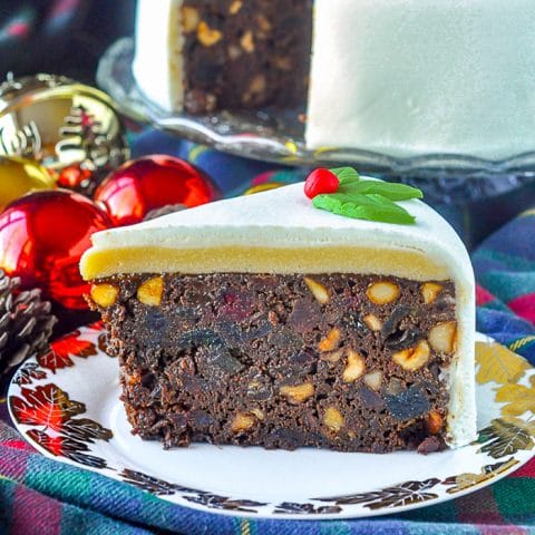 Ultimate Nut Fruitcake slioce on plate with gold leaf, Christmas tree ornaments in background.
