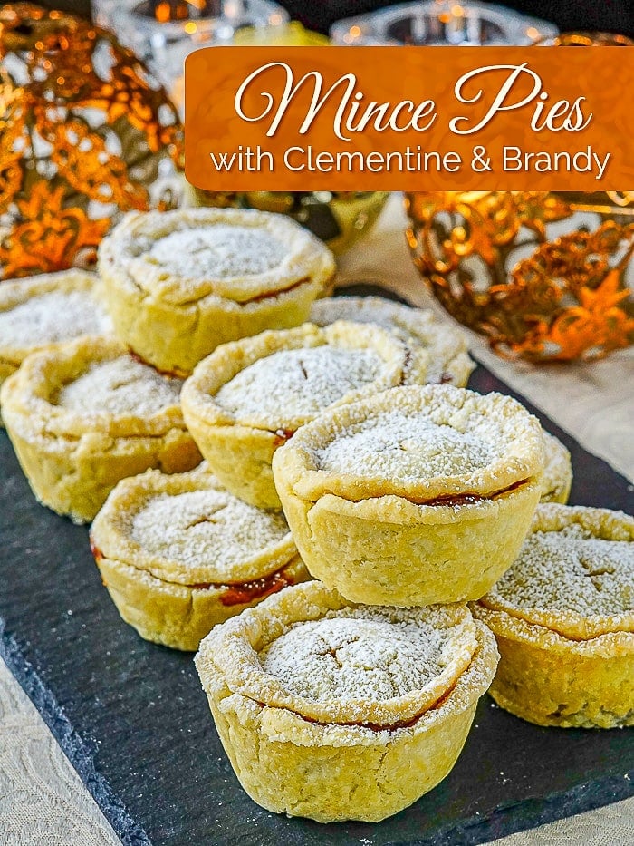 Mince Pies with clementine and brandy on a slate serving board with Christamas decorations in the background.