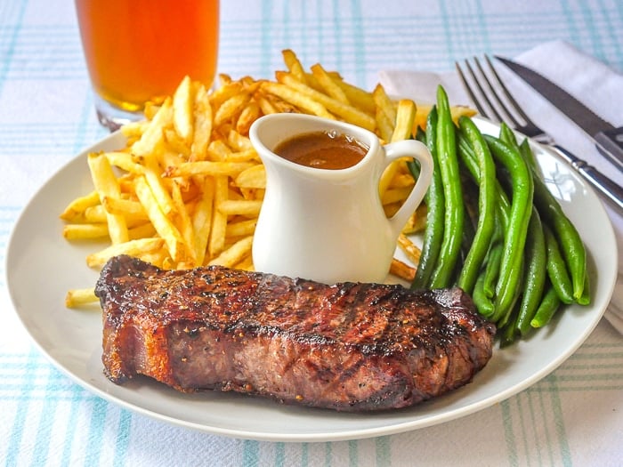 Steak Frites on a white plate with grren beans, gravy and a glass of beer in background