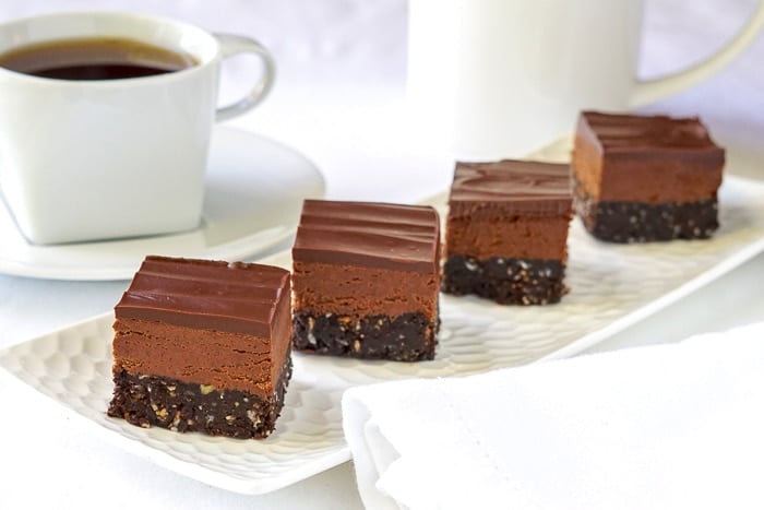 Chocolate Nanaimo Bars on a white platter with coffee in the background