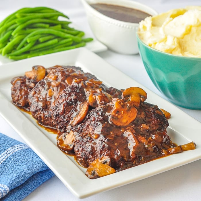 Salisbury Steak on a white serving platter with mashed potstoes, gravy and green bean s in the background