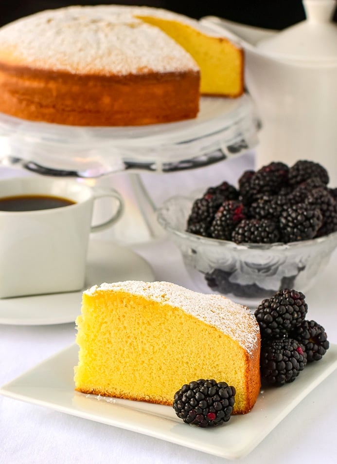 One slice of Condensed Milk Cake with blackberries coffee and a cake pedestal in the background