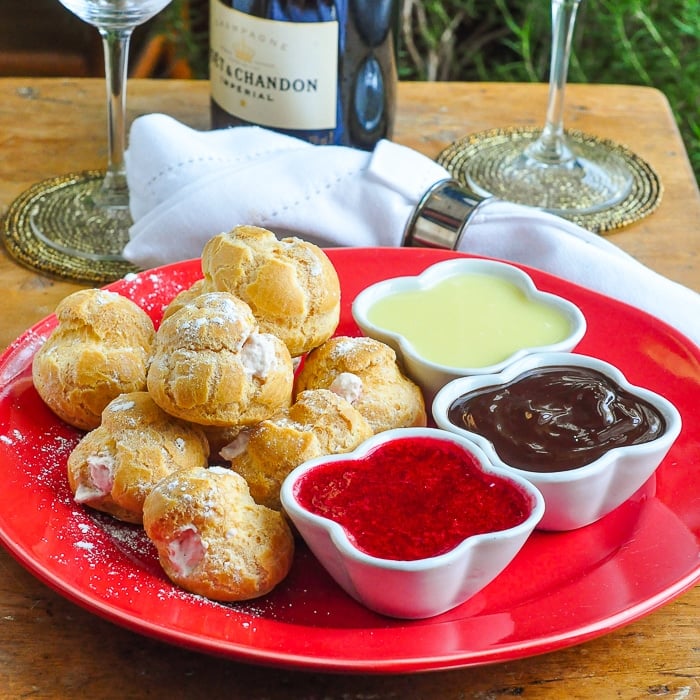 Raspberry Cream Profiteroles shown on a red plate with small ramekins of dipping sauces