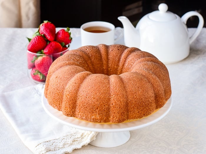 Photo of uncut Custard Bundt Cake on a white cake stand with strawberries and a tea service