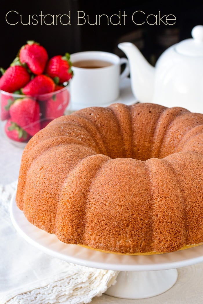 Photo of uncut Custard Bundt Cake on a white cake stand with strawberries and a tea service