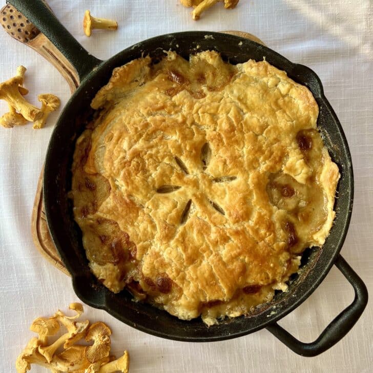 A chicken pot pie prepared in a skillet, on a white tablecloth with surrounding chanterelle mushrooms.