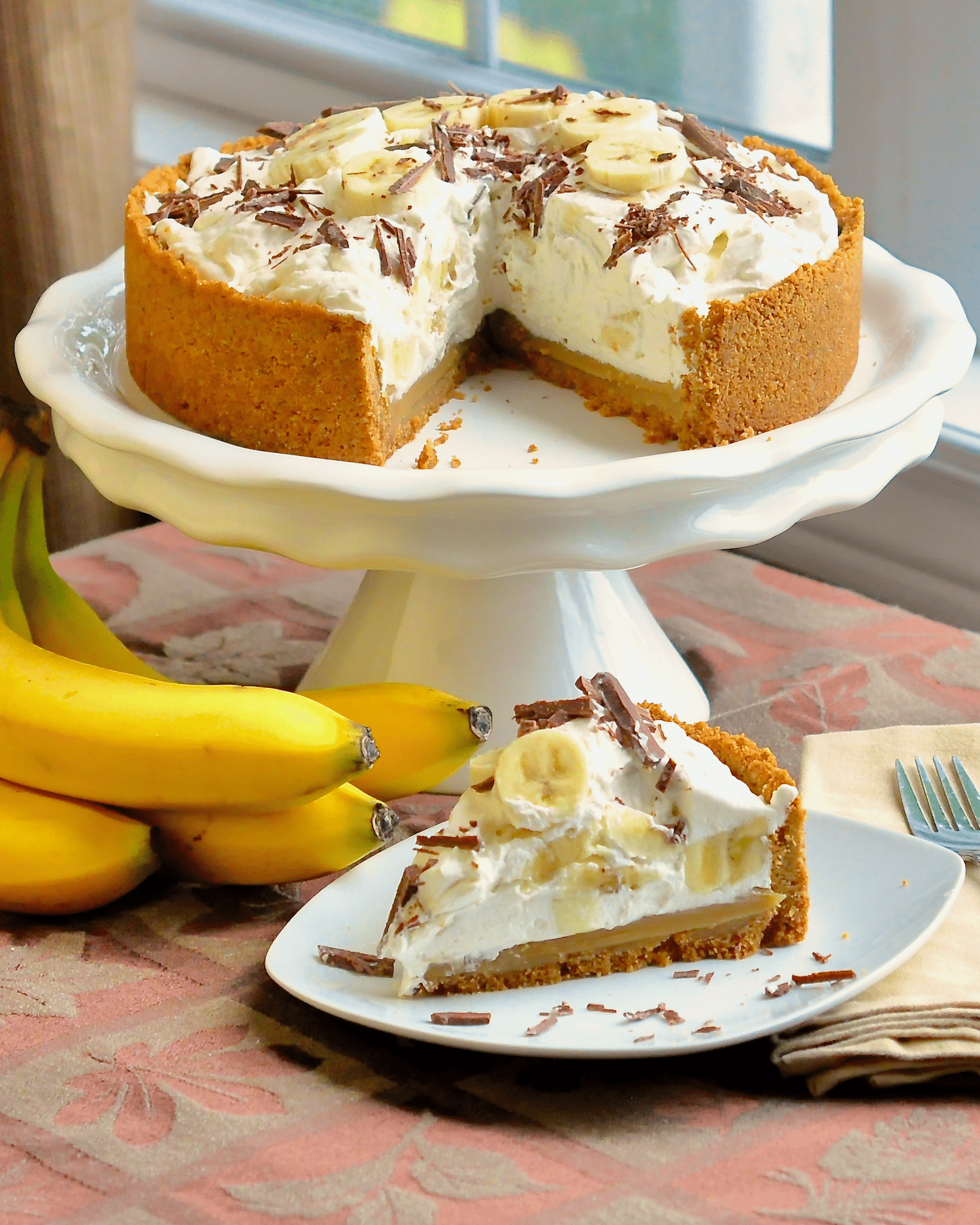 Photo of one slice of banoffee pie with whole pi eon background on a white pedestal
