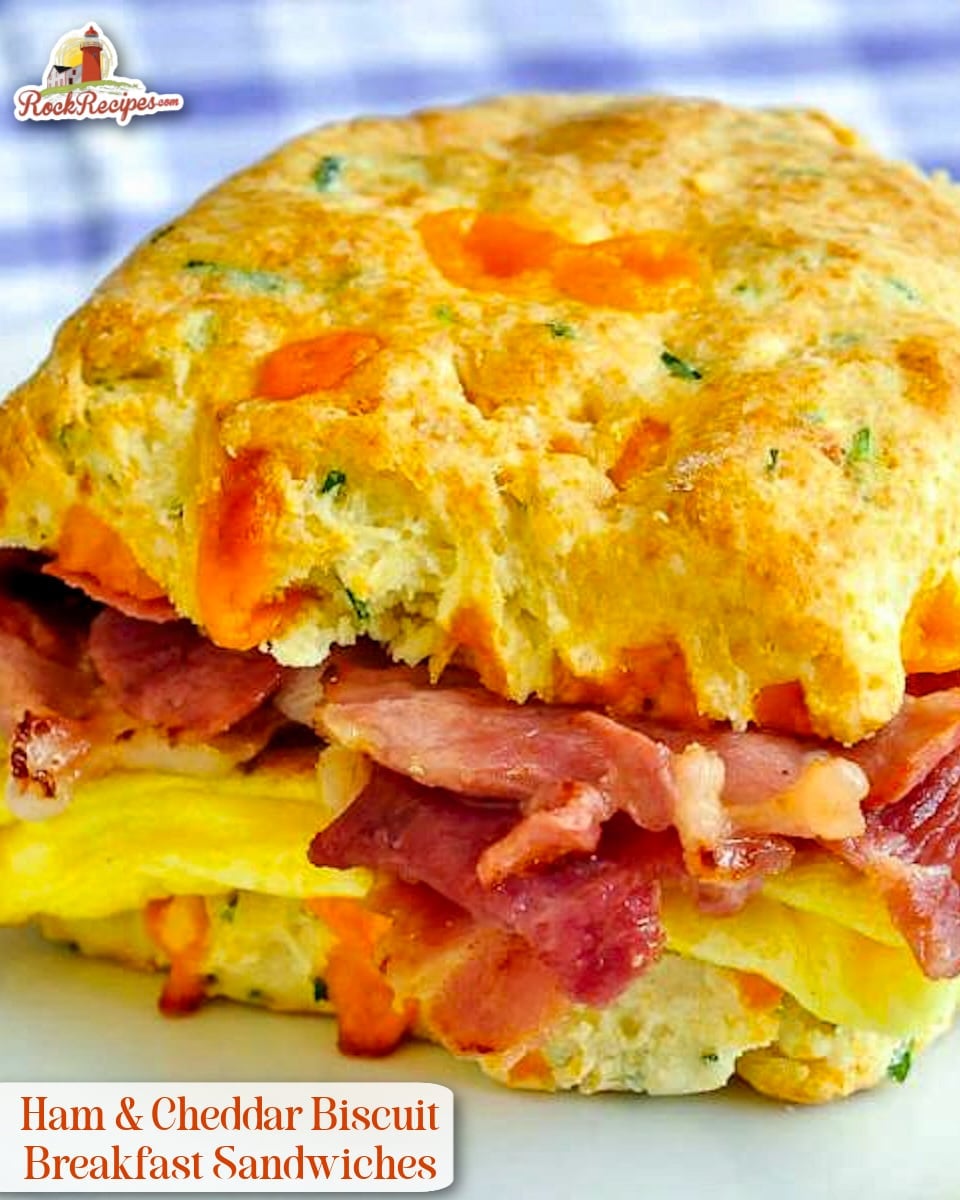 Ham Cheddar Biscuit Breakfast Sandwiches close up photo of one sandwich on a white plate on a blue checkered tablecloth with title text added for social media posts.
