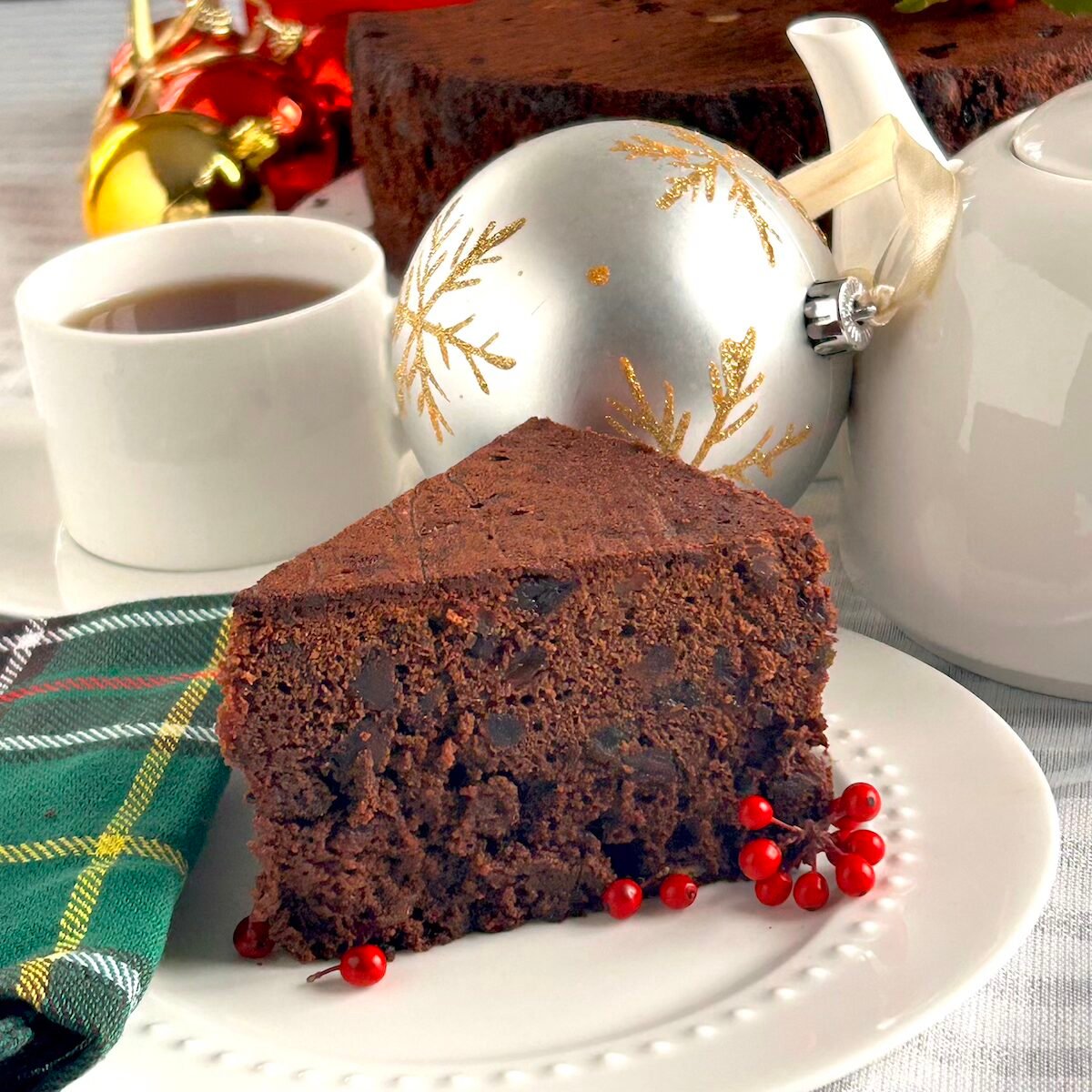 Chocolate Cherry Fruitcake close up photo of one slice on a white plate with Christmas decorations in background
