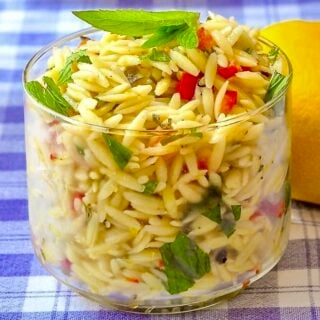 Close up photo of Lemon Mint Orzo Salad in a clear glass serving dish with a whole lemon on the side