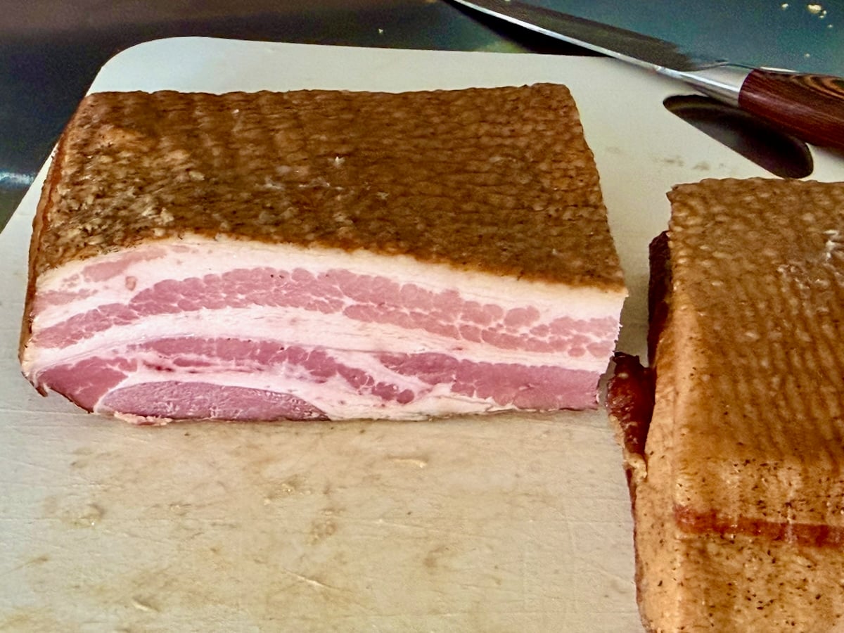 Photo of a slab of brown sugar cured homemade bacon being sliced on a white cutting board.