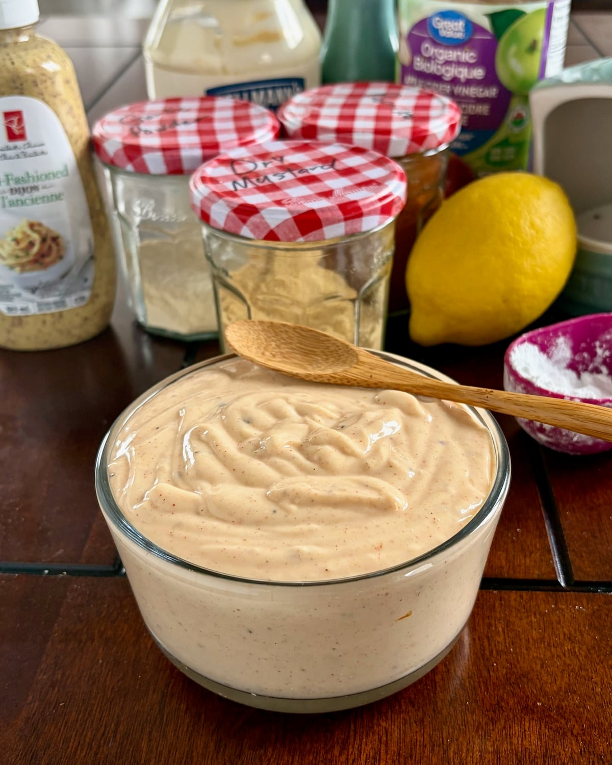 Photo of better than Miracle Whip salad dressing in a glass bowl with ingredients in the background and a wooden serving spoon on top.
