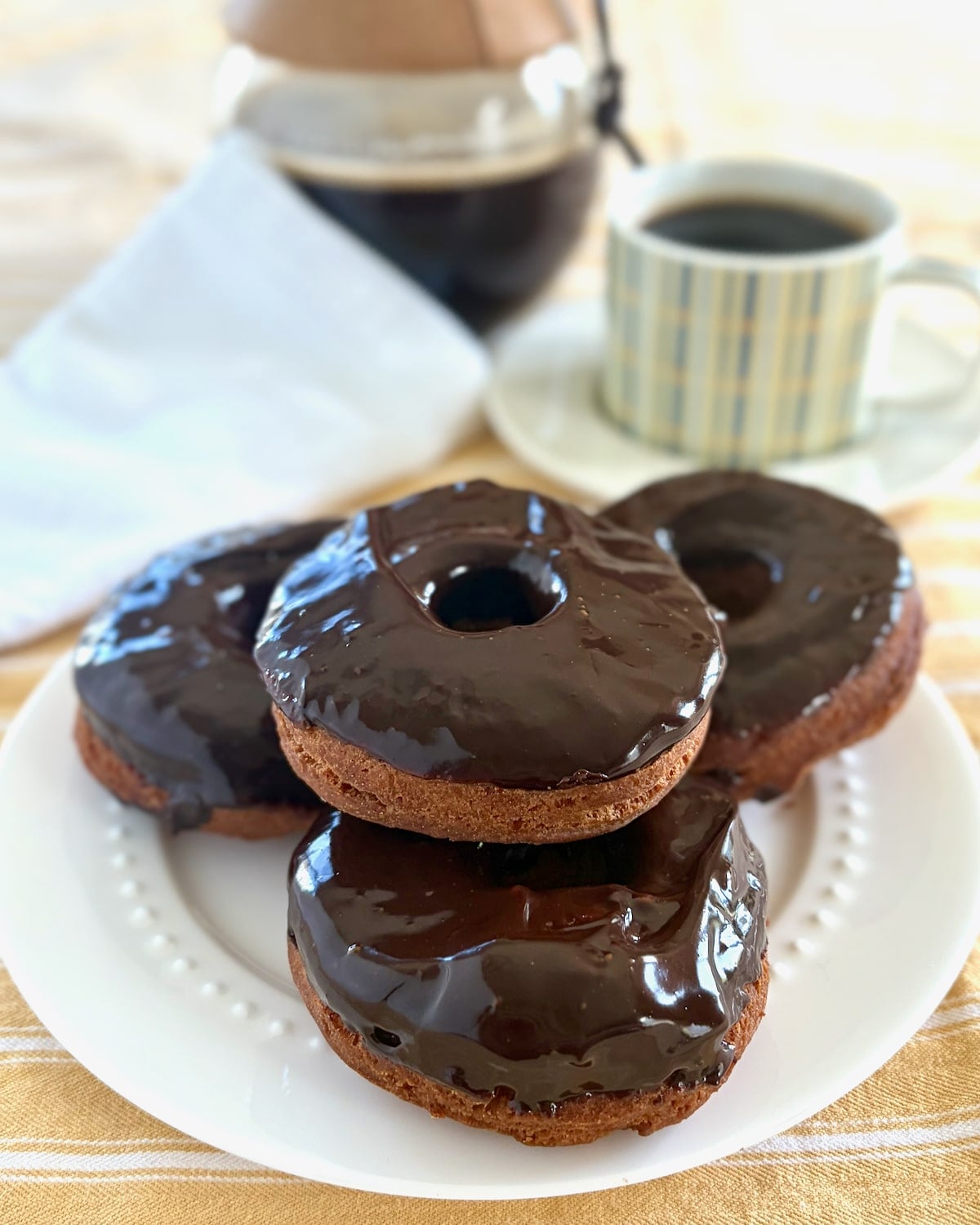 Photo of several chocolate glazed sour cream donuts on a white plate with coffee service in the background