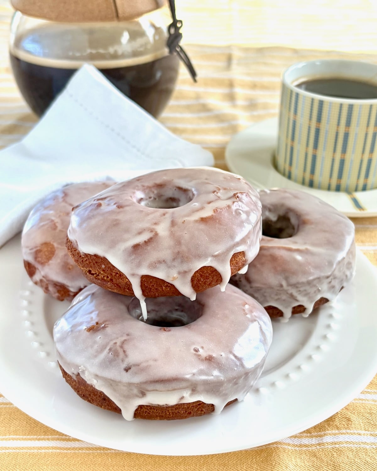 Rectangular cropped photo of vanilla glazed sour cream donuts stacked on a white plate with coffee service in the background.