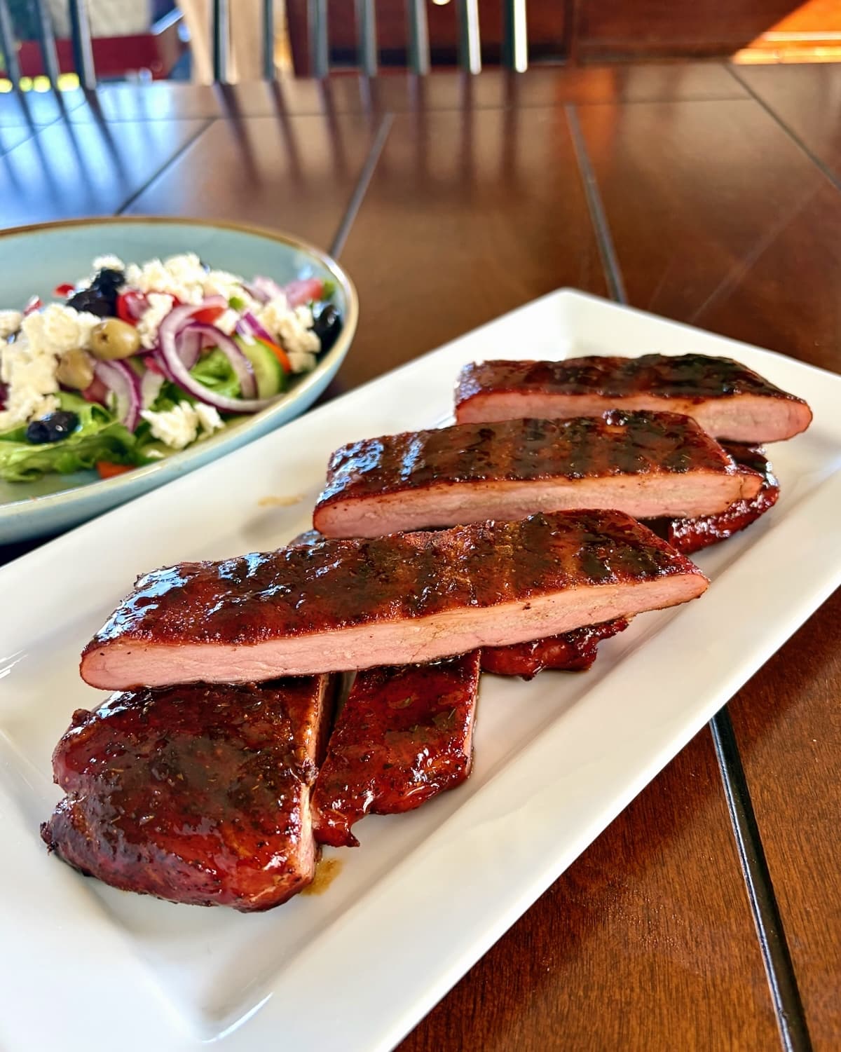 Barbecue Boneless Ribs sliced and stacked on a white serving platter, with greek salad in background.
