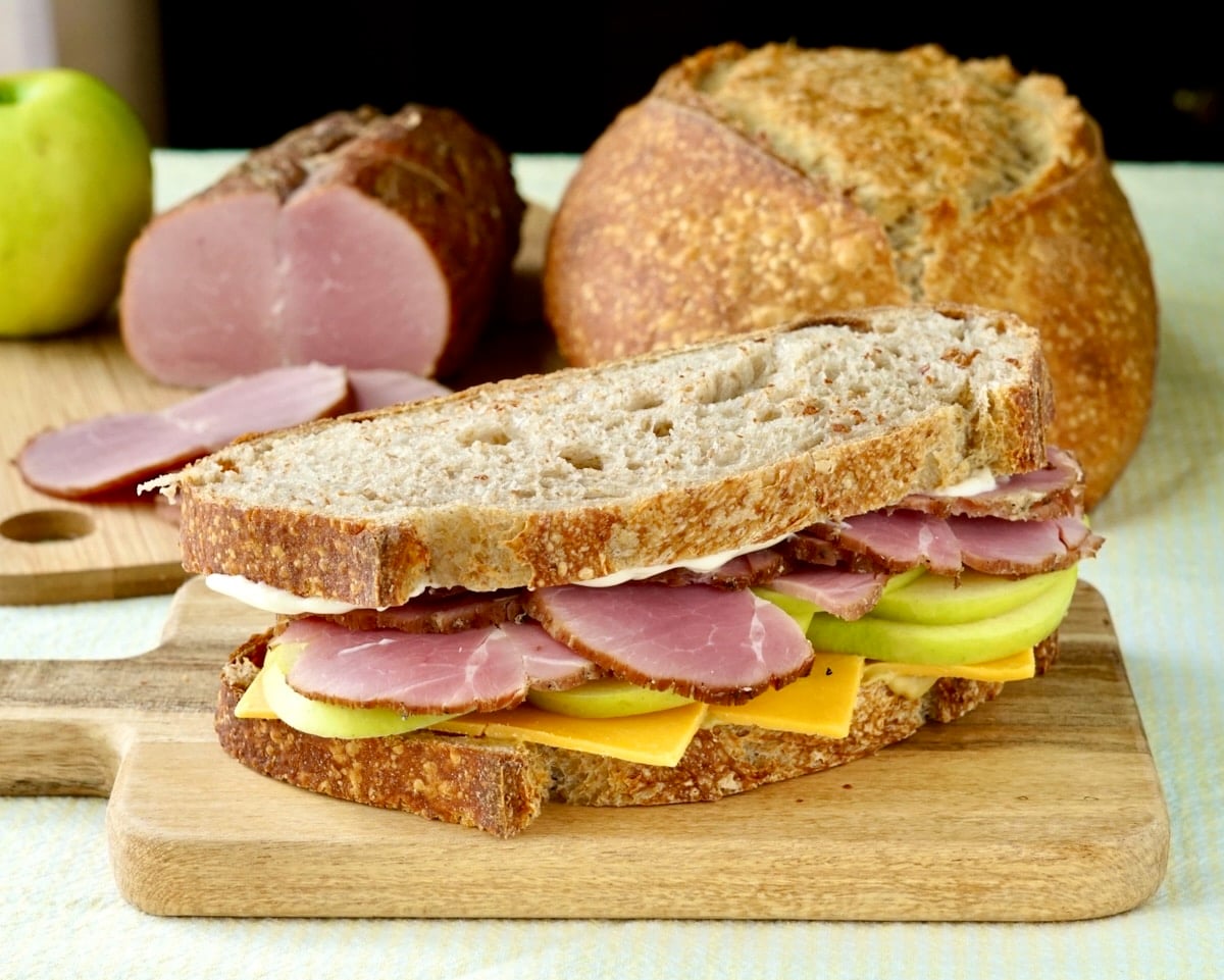 Wide shot photo of Photo of Homemade Tenderloin Ham on an apple cheddar and ham sandwich with sourdough bread in the background.