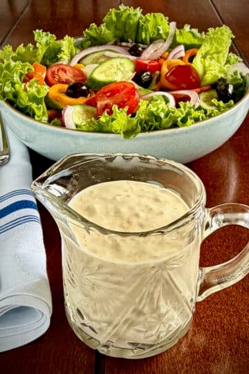 Close up photo of Creamy Lemon Parmesan Salad Dressing in a crystal jug dispenser with garden salad in the background,