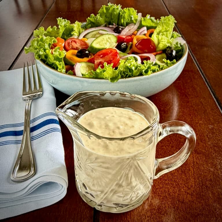 Close up photo of Creamy Lemon Parmesan Salad Dressing in a crystal jug dispenser with garden salad in the background,
