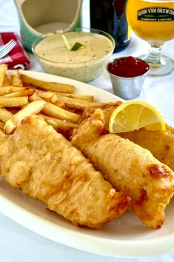 Fish and chips shown with tartar sauce and a glass of beer in the background.