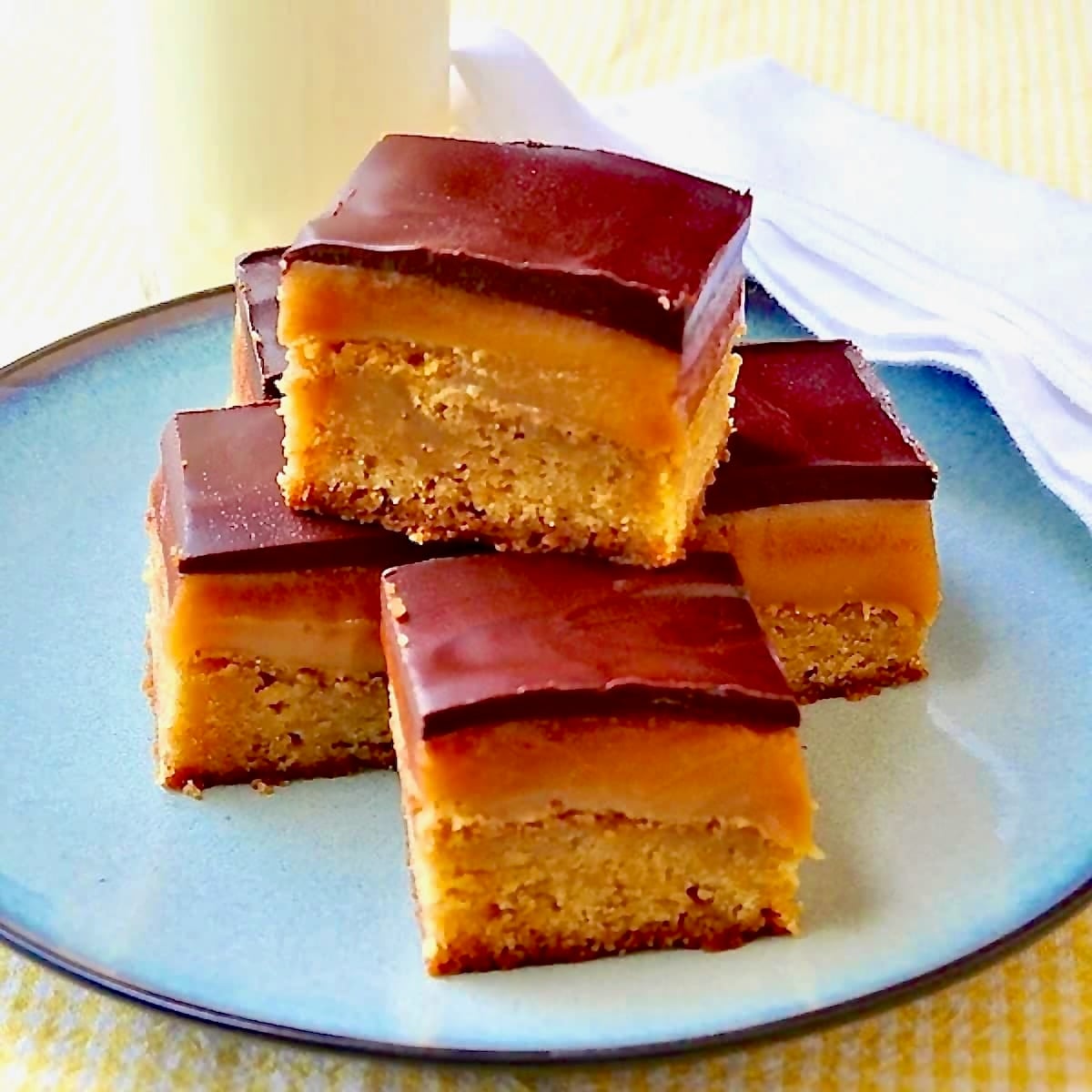 Millionaire Blondies photo of 5 cookie bars stacked on a blue plate with a glass of milk in the background.