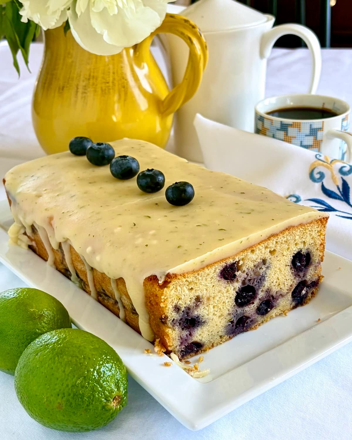 Photo of completed , uncut Glazed Blueberry Lime Loaf Cake on a white serving platter with one slice missing.
