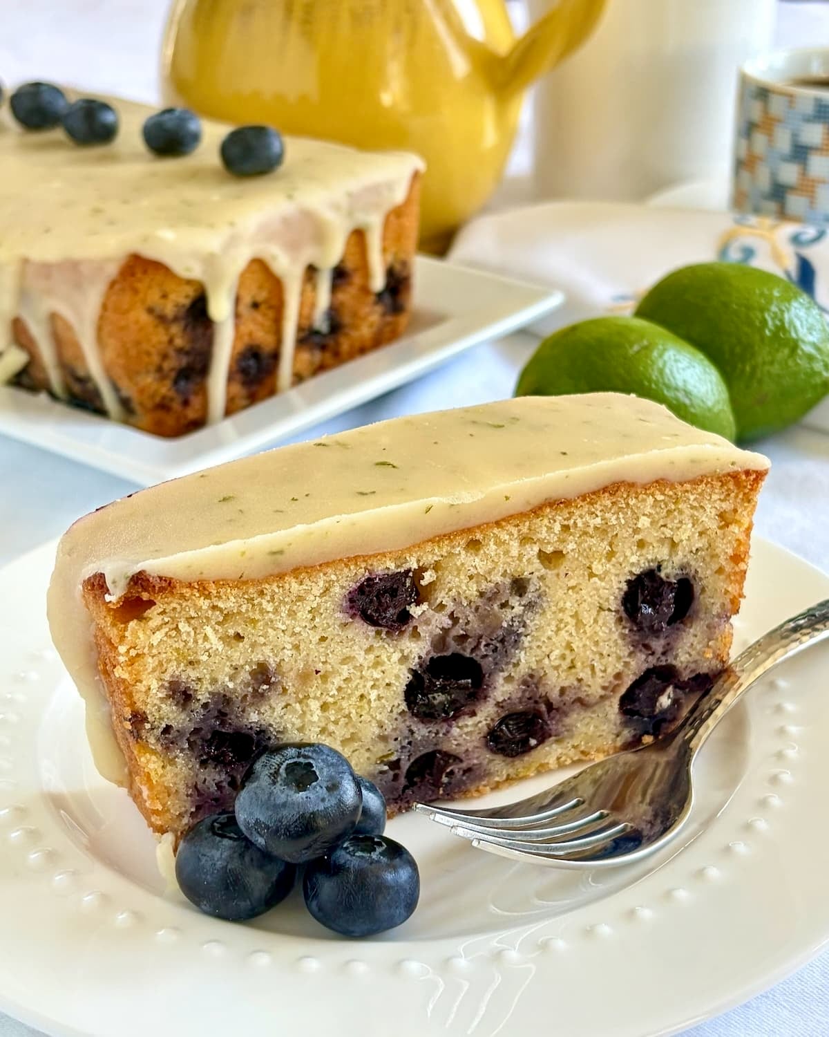 Close up photo of one slice of Glazed Blueberry Lime Loaf Cake on a white plate with blueberry garnish.