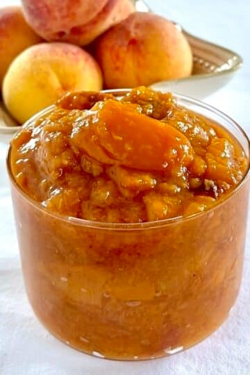 Easy Roasted Peach Jam pictured in a clear glass serving dish with fresh peaches in the background.