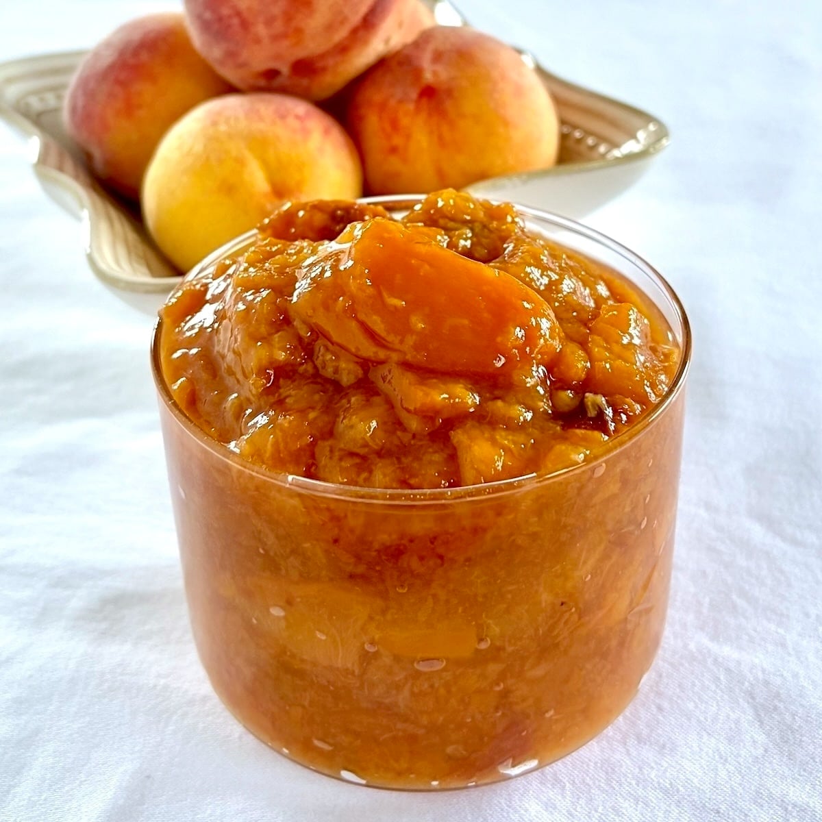 Easy Roasted Peach Jam pictured in a clear glass serving dish with fresh peaches in the background.