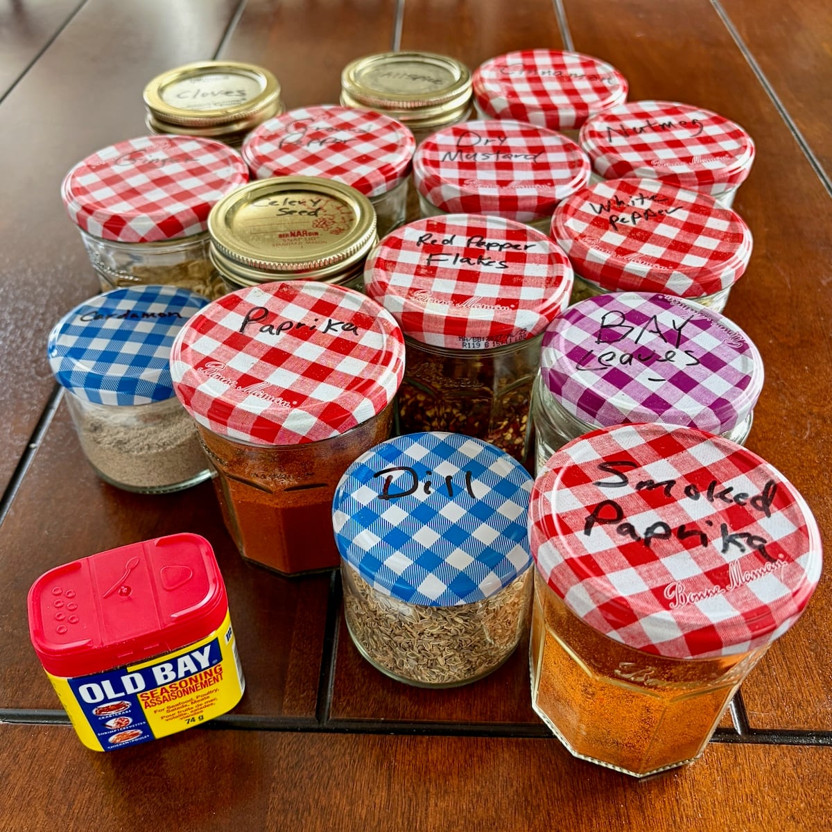 Photo of all the herbs and spices needed for Copycat Old Bay Seasoning in mason jars with a canister of Old Bay Seasoning in the foreground.