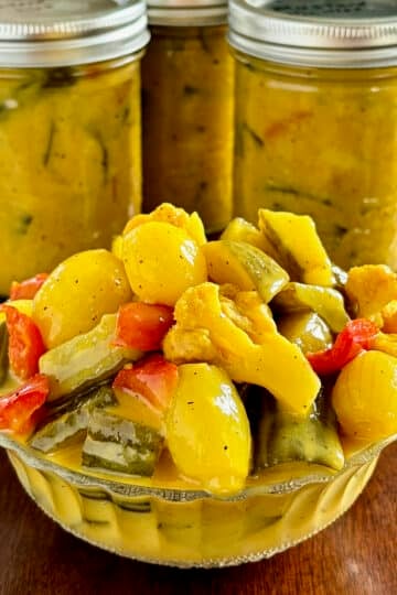 Newfoundland Mustard Pickles photo of mixed pickles in an antique clear glass serving dish with bottles of finished pickles in the background. Photo is cropped square as a Google Featured Image.